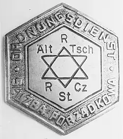 Insigne argentée pentagonale, avec en son centre une étoile de David, et sur son pourtour l’inscription allemande « Ordnungsdienst » et celle en polonais « Służba porządkowa ».