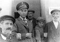 Un homme en veste de costume à rayures et cravate, coiffé d’un képi doté d’une étoile de David et portant un brassard sur lequel est lisible une partie du mot Ordnungsdienst pose face au photographe, dans l’embrasure d’une porte, entouré de trois hommes plus âgés et d’un enfant.