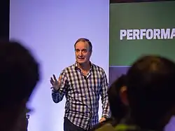 Un homme blanc entre-deux-âges fait une présentation, habillé d'une chemise à carreaux.