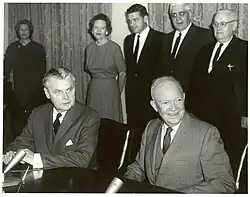 Diefenbaker and a smiling bald man in a suit sit at a table.  Two women and two men stand behind them.