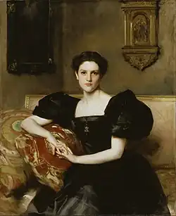 Portrait d'une femme assise, vêtue d'une élégante robe noire à manches bouffantes, dans un décor intérieur raffiné. Elle repose son bras sur un coussin richement orné de motifs rouges et dorés. Son regard est direct et confiant, et elle porte un collier avec un pendentif en croix. Son expression, à la fois sobre et intense, est accentuée par le jeu de lumière qui illumine son visage et ses mains. L'arrière-plan présente des éléments décoratifs, dont un cadre sombre et une petite niche dorée avec une sculpture.