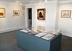 Galerie lumineuse avec œuvres encadrées et vitrine d'exposition. Palette neutre de blancs et crèmes, vitrine bleu sarcelle-gris. Murs, vitrine et sol lisses, textures réalistes, éclairage uniforme.