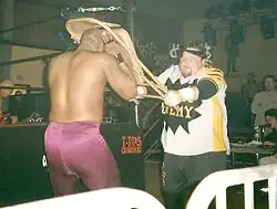 Catcheur blanc habillé d'un t-shirt blanc siglé des mots «&nbsp;Public Enemy&nbsp;» en jaune sur une étoile noire, d'un pantalon de catch noir et d'un bandana roulé noir sur la tête, portant un coup de chaise pliante sur la tête d'un autre catcheur noir portant un collant de catch violet. Ils sont au pied du ring. Un catcheur est couché dans le ring. On aperçoit un commentateur et l'arbitre à l'arrière.