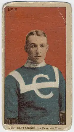 Portrait d'un jeune homme portant un chandail bleu avec col blanc, décoré par un sigle formé de la lettre C, en blanc sur la poitrine, traversée horizontalement par une bande blanche qui tourne vers le haut, à la gauche et à la droite de l'avant du chandail, et passe par-dessus les deux épaules.