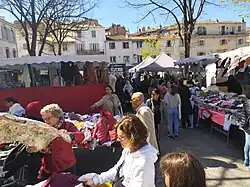 Le marché des forains fait partie de l'identité de la place - Des gens font leurs courses, marchent, discutent, observent des habits. La scène se déroule entre plusieurs stands de vêtements et de fripes. On aperçoit un stand de fruits et de légumes à l'arrière plan.