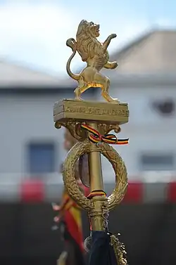 Fleuron de hampe de drapeau belge avec couronne de feuilles de chêne et de laurier, la devise nationale et le lion héraldique.