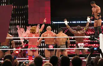 À droite, Justin Gabriel se tient debout sur la troisième corde du ring. Il s'apprête à effectuer sa prise de finition, le 450e splash, en se jetant sur Ricky Steamboat (non visible sur l'image). Les autres membres du Nexus, de dos, l'encouragent.