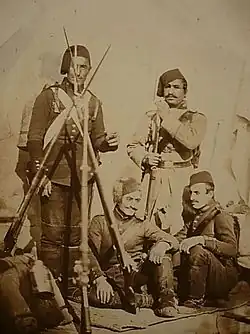 Photographie de quatre soldats moustachus portant des fez. Deux sont assis à côté d'un trépied de fusils équipés de baïonnettes.