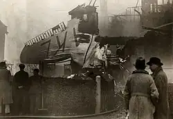 Photo noir et blanc de débris d'avion sur une maison en ruine, avec des badauds en train de discuter au premier plan.