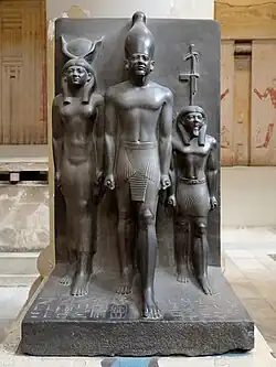 Triade du 4e&nbsp;nome de Haute-Égypte, Thèbes, nome du Sceptre. Le personnage du nome est un homme très petit. 93&nbsp;×&nbsp;47&nbsp;cm. Musée du Caire JE 40678