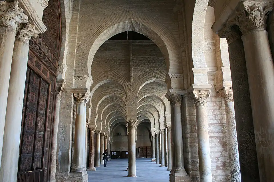 Vue intérieure montrant une double rangée d'arcades en perspective dans le portique précédant la salle de prière. Les arcs sont de type outrepassé (brisé ou en plein cintre).