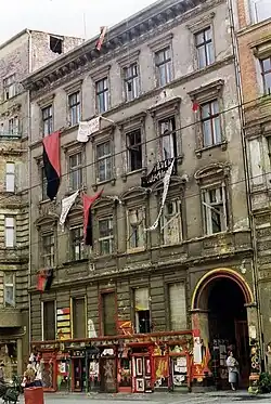 Photo couleur d'un immeuble. Sont suspendus aux fenêtres plusieurs drapeaux et banderoles, dont deux rouge et noir.