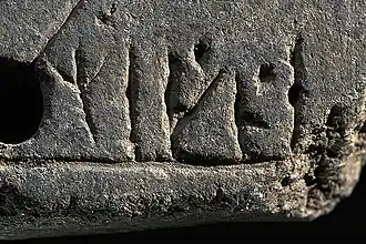 Photographie en couleurs d'une pierre rectangulaire gris foncé marquée de graphèmes, la partie gauche percée d'un trou, le coin en bas à droite présentant des stigmates de cassure.