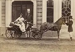 Assis dans une calèche dont le cheval est tenu en mains par un piqueur, Louise et son frère habillés de clair et coiffés d'un chapeau orné posent sous le regard de la future reine Marie-Henriette en tenue luxueuse claire à damiers foncés et du futur roi Léopold II vêtu d'une redingote sombre et d'un pantalon clair et coiffé d'un chapeau haut de forme
