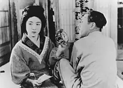 Une femme à l'air renfrogné montre ostensiblement un réveil à un homme assis à ses côtés et qui fait la moue (image en noir et blanc).