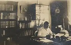 Photographie en noir et blanc datant de 1919, représentant Kirstine Smith assise à un bureau, habillée de façon formelle avec une blouse ou une chemise claire et une veste sombre. Ses cheveux sont attachés en un chignon bas, typique de la mode féminine du début du XXe siècle. Le bureau devant elle est sobre, avec des documents ou des papiers visibles, indiquant une activité intellectuelle ou académique. Elle a une expression sérieuse et concentrée, son regard tourné légèrement vers la droite, ce qui suggère qu'elle est en train de réfléchir ou de travailler. Le cadre de la photo est simple, sans éléments décoratifs notables, centrant l’attention sur Kirstine Smith elle-même. L’éclairage est uniforme, sans ombres marquées, ce qui est caractéristique des portraits photographiques de cette époque.Cette photo reflète son engagement dans le domaine scientifique à une époque où les femmes étaient encore peu présentes dans ce milieu. La photographie est issue d'une collection privée et son auteur est inconnu. Elle est désormais dans le domaine public, conformément à la législation danoise sur le droit d'auteur.