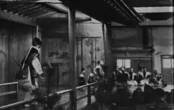 Premier plan à gauche : l'acteur s'avance sur le pont ; à droite, les spectateurs vus de dos. Au fond, musiciens accroupis.