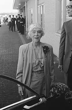 souriante, Élisabeth vêtue d'un tailleur clair pose près d'une voiture devant les bâtiments d'un aéroport