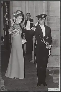 Image montrant un couple debout composé d'une jeune femme qui salue de la main droite et d'un homme en tenue d'officier de marine faisant le salut militaire.
