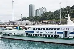 Photographie d'un ferry sur lequel il est écrit « Labuan Express 2 ». À l'arrière-plan se trouvent deux gratte-ciel.