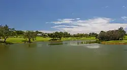 Photographie d'une partie d'un terrain de golf qui présente une grande étendue d'eau et une dense végétation tropicale.