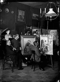 Photographie de Léon Philippet, de dos, en train de peindre une toile sur un chevalet devant deux hommes à sa gauche qui l'observent travailler. On aperçoit trois autres toiles de l'artiste à côté ou derrière celle sur laquelle travaille l'artiste.