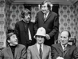 Photo en noir et blanc de cinq hommes en costumes faisant les pitres dans un salon.