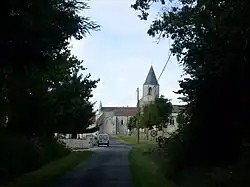Une petite route bordée d'arbres avec au fond une église de pierre couverte de tuiles au clocher conique ardoisé. Un panneau de limitation de vitesse à 30 km/h à droite.