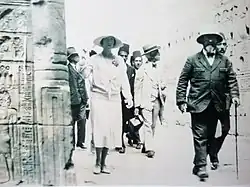 vêtue de clair, la reine chemine en compagnie du professeur Capart dans une allée d'un temple dont on aperçoit à gauche l'un des murs gravés de hiéroglyphes