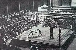 Photographie montrant deux hommes luttant et un juge sur un ring au milieu des spectateurs, depuis un point de vue en hauteur.