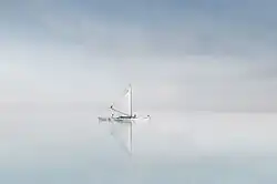 Une athmosphère cotoneuse, composée d'eau et de ciel, tout est calme et plat. Au centre, un petit bateau, avec son reflet estompé, une voile blanche pointée vers le ciel.