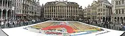 Vue panoramique d'une planche géante montrant une fusée sur sa tour de montage, au centre de la Grand Place de Bruxelles, les spectateurs étant séparés de l'image par des barrières.