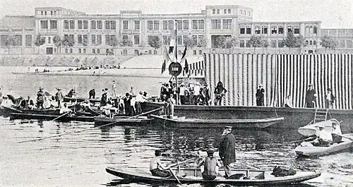 Photographie noir et blanc : des barques sur l'eau autour d'un ponton encombré de nombreuses personnes.