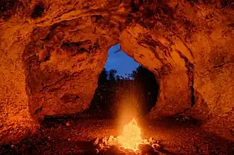 Photographie en couleurs d'un feu consumant des bûches en bois, sa lumière rougeoyante se réfléchissant sur les parois d'une grotte.