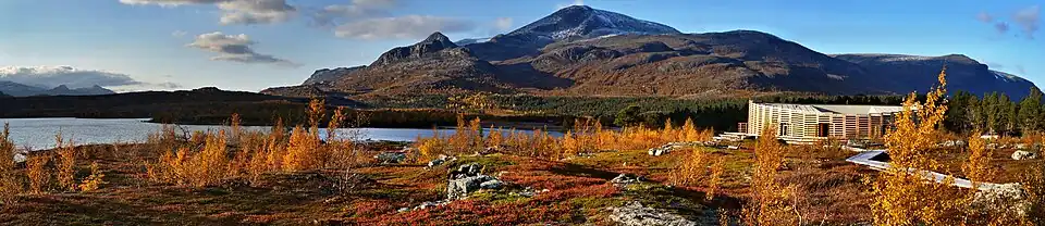 Un bâtiment dans une forêt en automne, près d'un lac, avec des montagnes en arrière-plan.