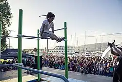 Photo d'une femme en position d'équerre sur une barre horizontale. Du public l'observe en contre-bas.