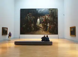 Vue générale d'une salle de musée avec un immense tableau au fond et deux autres sur les murs latéraux. Au premier plan, une banquette où sont assis deux personnages