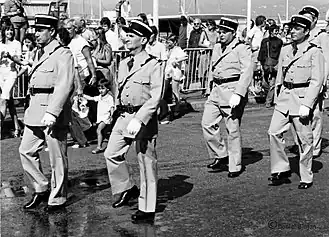 Photo en noir et blanc montrant six gendarmes en uniforme d'apparat défilant sous les regards de touristes.