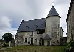 Photographie en couleurs d'un bâtiment à toit pentu accolé d'une galerie en hauteur et d'une tourelle octogonale, la façade d'un deuxième bâtiment visible sur la droite, un arbre s'élevant sur la gauche, une étendue herbeuse apparaissant au premier plan.