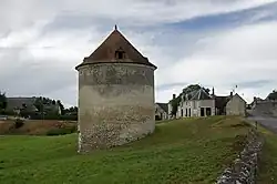 Photographie en couleurs d'un bâtiment circulaire à toiture pentue de tuiles s'élevant au milieu d'un pré, un muret en pierre visible sur la droite, des constructions anciennes et des maisons apparaissant en arrière-plan