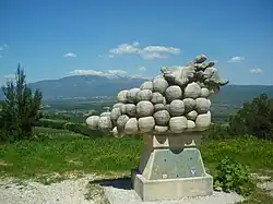 La photo en couleur représente une sculpture de grande taille d'une grappe de raisin. Elle est juchée sur un socle. Au second plan, dans un vallon, des vignes marquent de leur damier vert alors qu'en arrière-plan, le mont Ventoux domine le paysage.