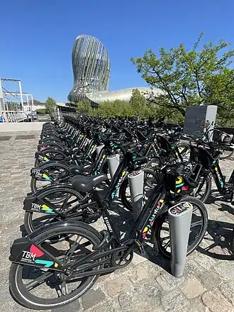 Photographie d'une station de Le Vélo. Les vélos noirs sont alignés devant l'imposante cité du vin.