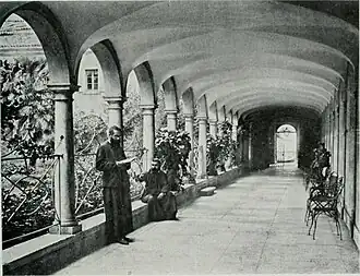 Photographie en noir et blanc montrant une galerie avec des arcades séparant d'un jardin. Deux moines sont en train de lire, l'un debout, l'autre assis.