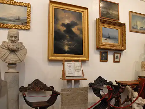 Photographie couleur d'un mur sur lequel sont accrochées plusieurs peintures à l'huile. En bas à gauche se trouve le buste sculpté d'un homme barbu et au centre une plaque commémorative en marbre.