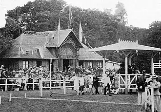 Image d'un bâtiment devant une forêt, des personnes sont le long d'une piste d'herbe.
