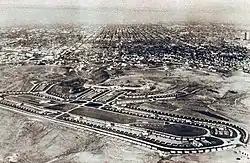 Image en noir et blanc. Au fond, la ville de Los Angeles. À l'avant-plan, un immense ovale bordé de petites maisons de plain-pied, qui forment le village olympique ; l'intérieur de l'ovale contient trois bâtiments en longueur et de la verdure.