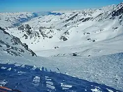 Photographie d'un glacier dans un vallon, avec des sommets enneigés au fond de la photo, le paysage étant enneigé : au centre, la neige est partout hormis sur de hauts rochers noirs qui dépassent ça et là avec un peu d'ombre à gauche de la photo où la montagne du Peyron s'assombrit.