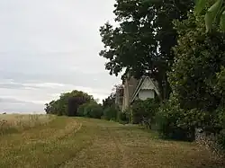 Photographie en couleurs d'un champs bordé par des haies et la clôture d'un manoir