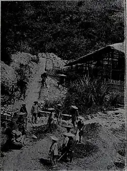Photographie en plan éloigné, montrant un paysage où l'on aperçoit des hommes, coiffés de « chapeaux chinois », en train de travailler.