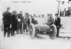 Photographie en noir et blanc d'une ancienne voiture de course, avec de nombreuses personnes à côté.